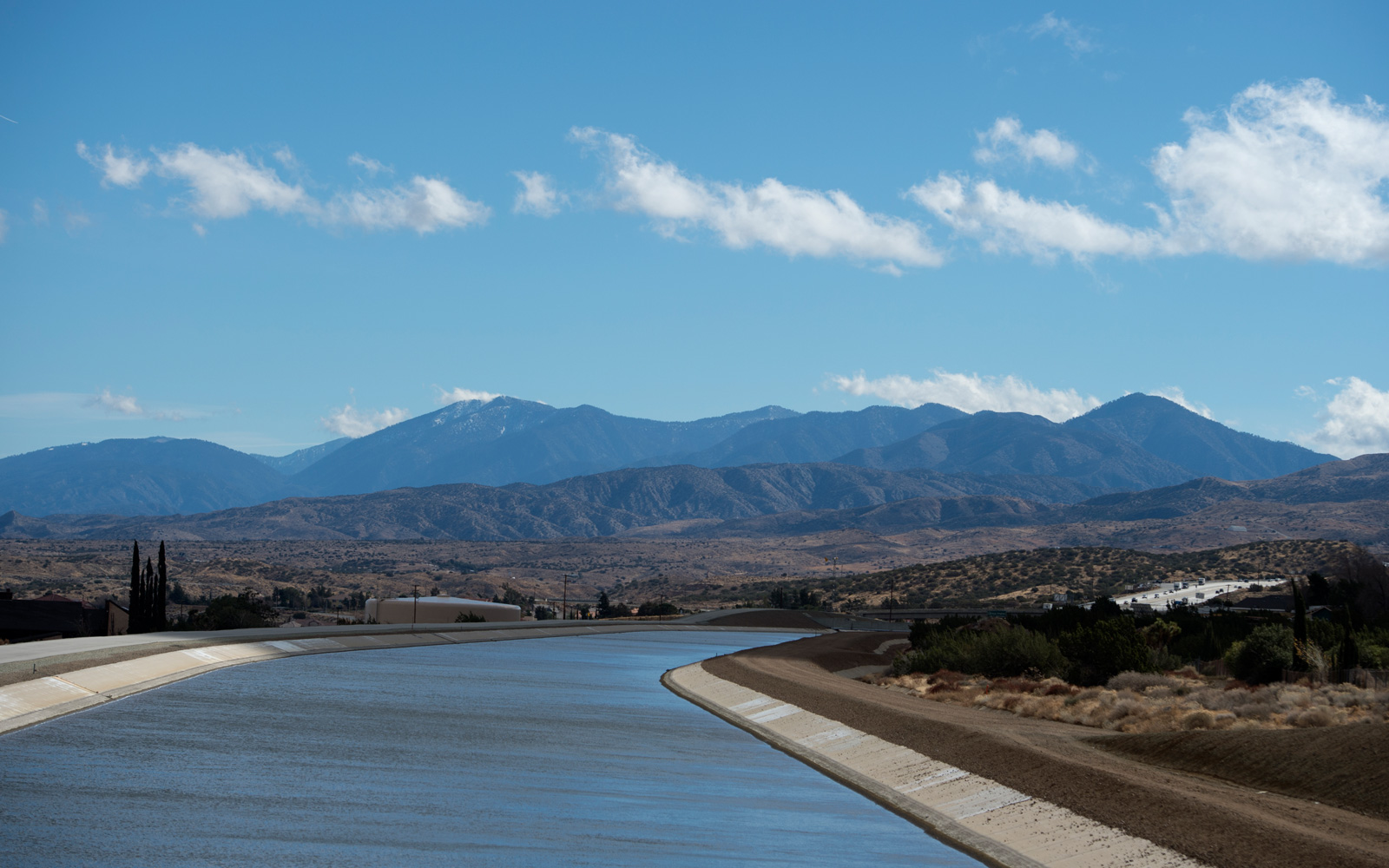 A long, concrete water aqueduct channel stretching through a dry, mountainous landscape under a bright blue sky with scattered clouds