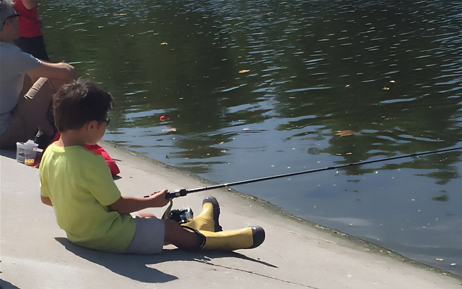 A young child wearing a yellow shirt and boots sits by the edge of a calm lake holding a fishing pole.