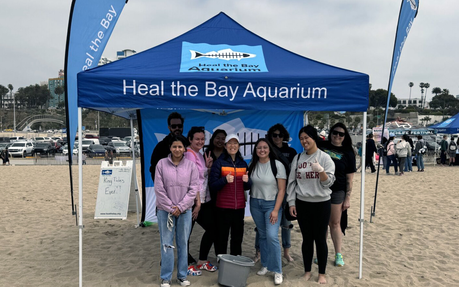 A group of volunteers posing in front of a Heal the Bay Aquarium tent on a sandy beach
