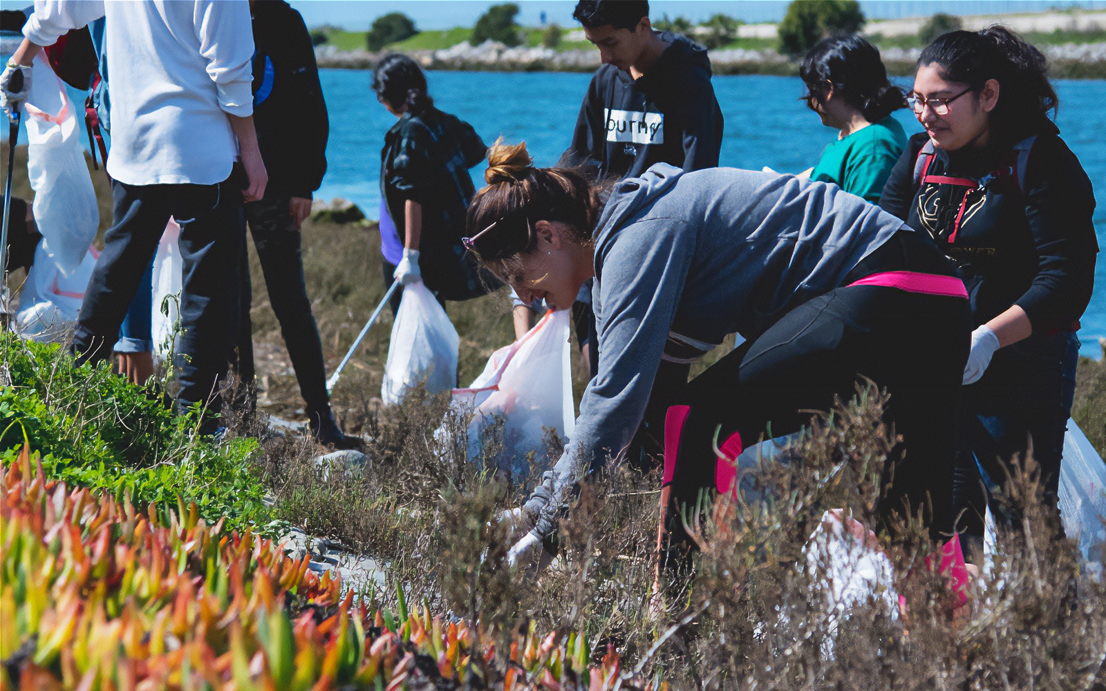 A group of volunteers and students gathering litter and debris along a riverbank during a community cleanup event