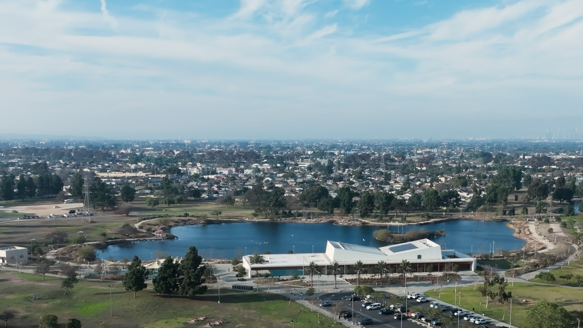 An aerial view of the Earvin “Magic” Johnson Recreation Area featuring a large lake, modern community buildings, and walking paths surrounded by urban residential neighborhoods.