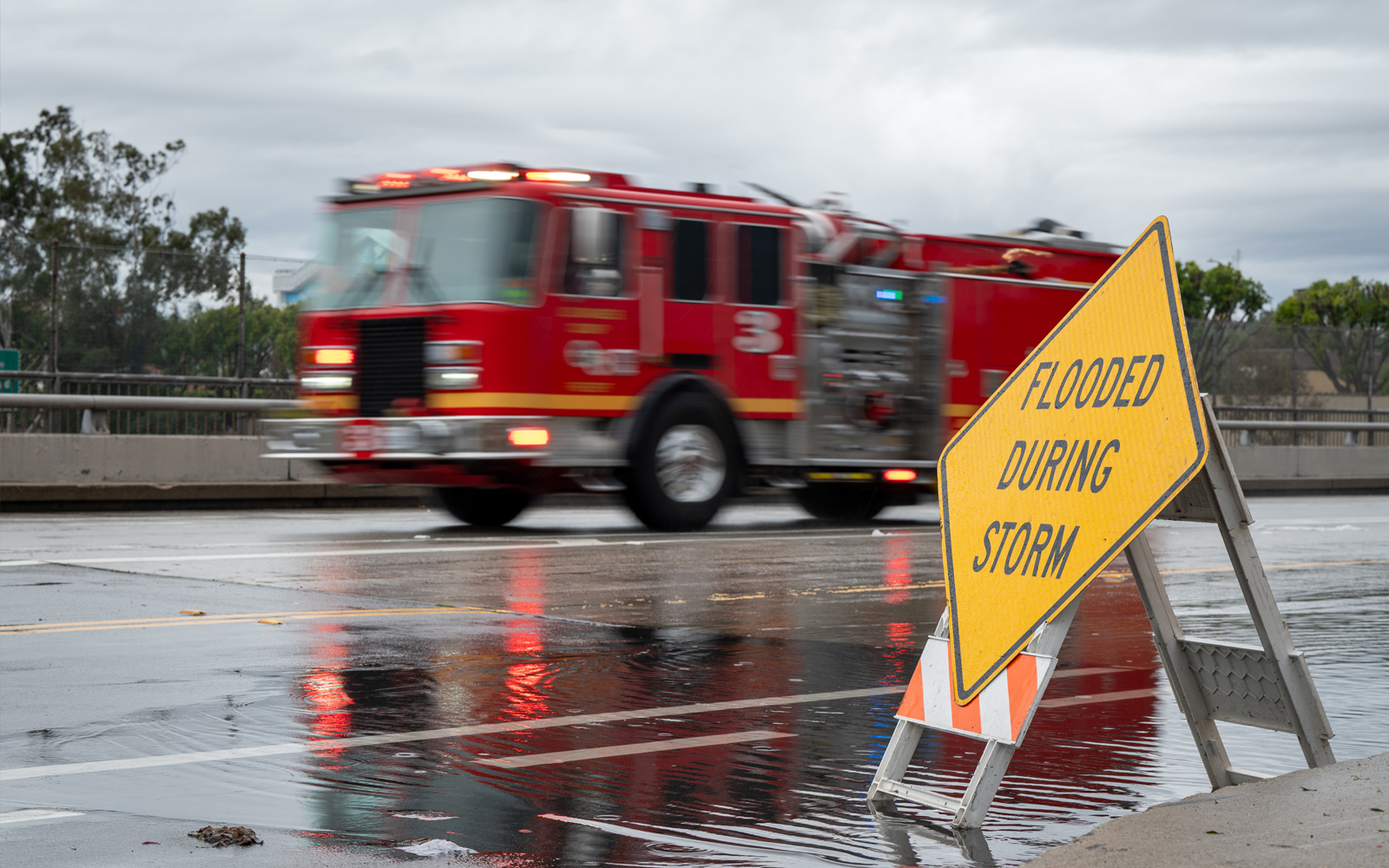 A blurred fire truck driving past a yellow "Flooded During Storm" warning sign on a wet city street